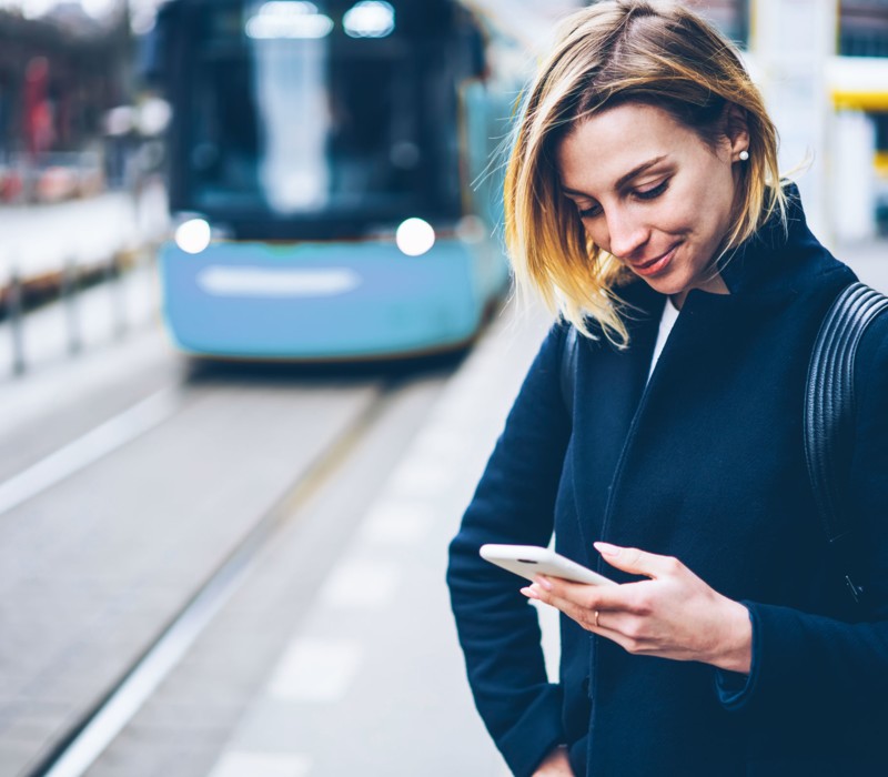 Une jeune femme en manteau noir au bord de la route regarde son téléphone en souriant, tandis qu’un tramway jaune s’approche à l’arrière-plan.