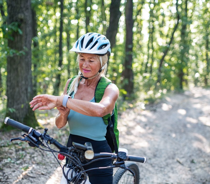 Eine blonde Frau mit Helm steht mit dem Fahrrad auf einer Waldstrasse und schaut auf ihre Sportuhr.