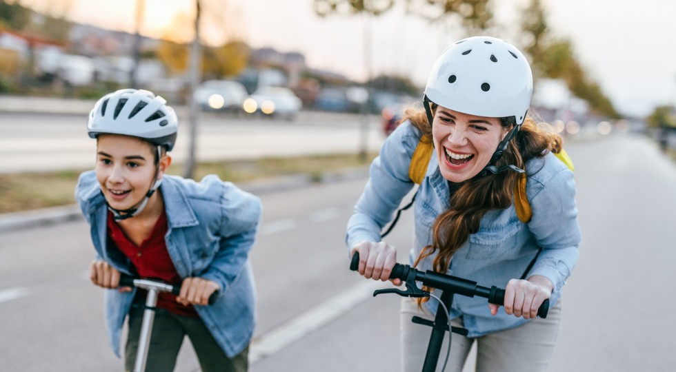 Zwei Jugendliche mit Helm und Jeanshemden fahren lachend auf E-Scootern auf einer breiten Strasse.