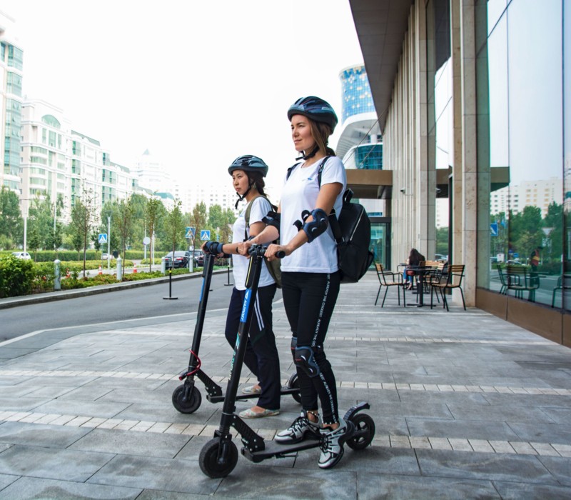 Zwei Frauen stehen mit Helm und Gelenkschützen auf einem E-Scooter, bereit zur Abfahrt.