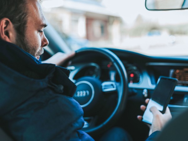 Ein Mann sitzt im Auto am Steuer und schaut auf sein Handy, das er in der rechten Hand hält.