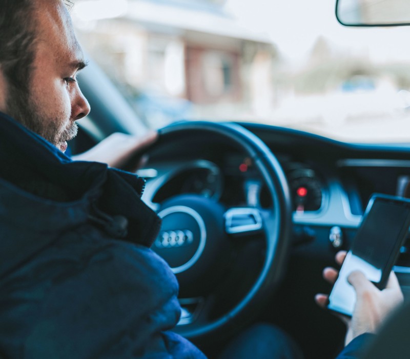 Ein Mann sitzt im Auto am Steuer und schaut auf sein Handy, das er in der rechten Hand hält.