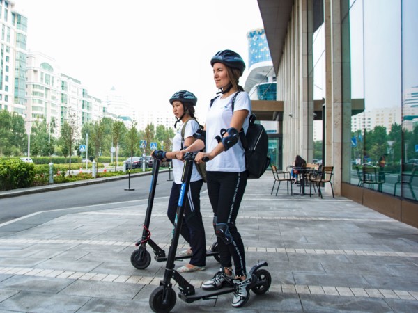 Deux femmes, équipées d’un casque et de protections articulaires, se tiennent prêtes à partir sur un scooter électrique.