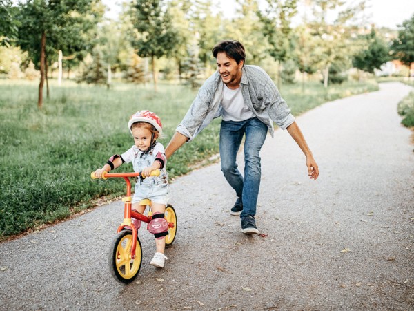 Un homme soutient une petite fille portant un casque et faisant du vélo sur une route verte dans un parc.