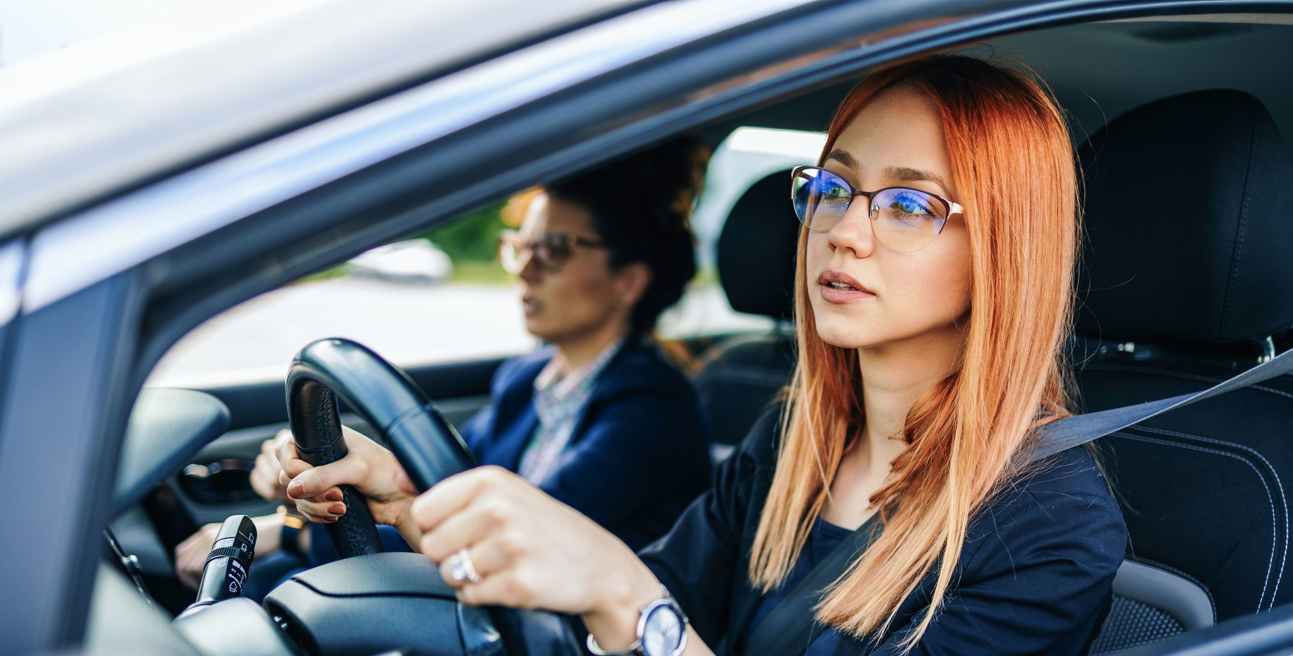 Une jeune femme rousse à lunettes est installée au volant et regarde la route attentivement, tandis qu’une passagère plus âgée est assise à côté d’elle.
