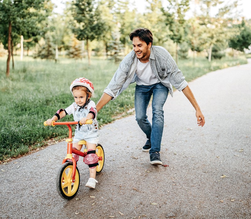 Ein Mann unterstützt ein kleines Mädchen mit Helm, das auf einer grünen Strasse im Park Fahrrad fährt.