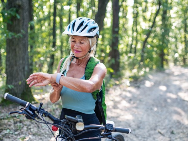 Une femme blonde portant un casque est avec son vélo sur une route de forêt et regarde sa montre de sport.